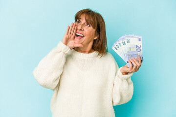 Middle age caucasian woman holding bank notes isolated on blue background shouting and holding palm near opened mouth.