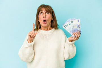 Middle age caucasian woman holding bank notes isolated on blue background pointing upside with opened mouth.