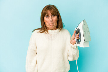 Middle age caucasian woman holding a iron isolated on blue background shrugs shoulders and open...