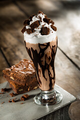 Chocolate milkshake with whipped cream and brownie pieces on top, on a wooden table