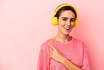 Young caucasian woman listening to music isolated on pink background smiling and pointing aside, showing something at blank space.
