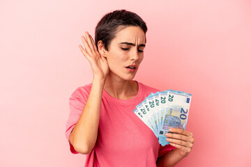 Young caucasian woman holding banknotes isolated on pink background trying to listening a gossip.
