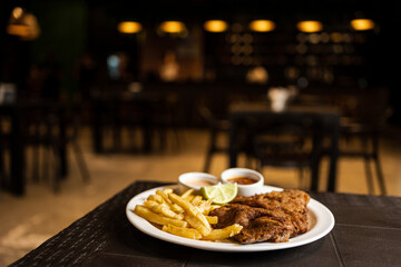 plate of milanesa (breaded meat) with french fries and lemon in a restaurant with a unfocused bar in the background