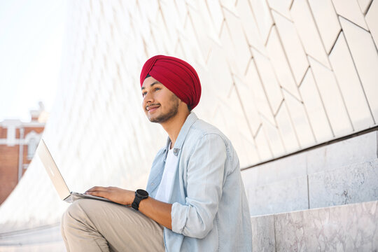 Optimistic Indian male student, employee, freelancer sitting outdoors and using laptop, successful young hindu entrepreneur in traditional turban headwrap with laptop sitting on the steps in cityscape