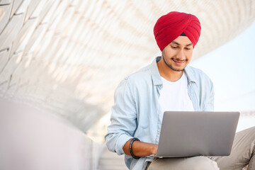 Indian freelancer man in traditional turban pagg sitting outdoors with laptop, developer working on the distance, hindu guy enjoys remote work, male student studying online, e-learning concept - obrazy, fototapety, plakaty