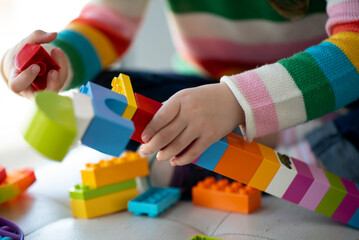 A cute little girl plays construction set. Childhood. Development. She sits near a window with a large teddy bear. The girl is dressed in a colored sweater and jeans.