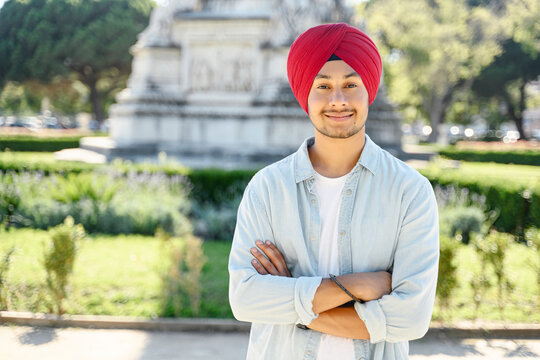 Handsome smiling young Indian man wearing traditional red turban stands outdoors with folded arms. Proud confident sikh man with arms crossed looking at camera standing in cityscape