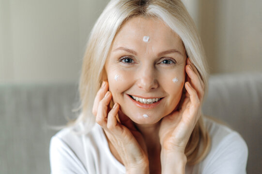 Mature blonde woman with cosmetic cream on face. Close-up of beautiful caucasian senior female, moisturizes the skin, applies anti aging cream, cares about facial skin, looks at camera, smiling