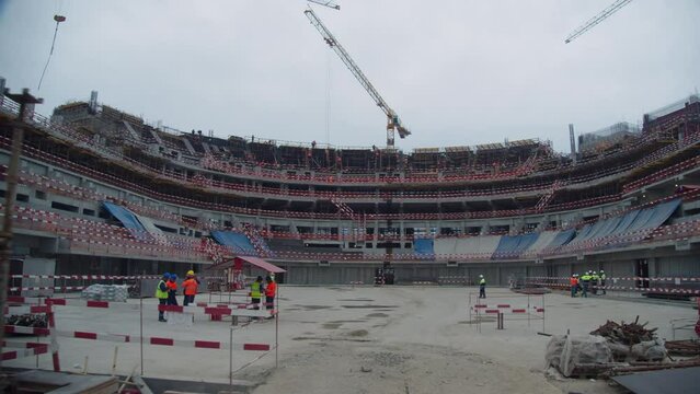 Foundation of modern hockey stadium building with working builders at construction site on cloudy day. City development
