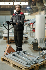 Young serious male worker of large warehouse standing by forklift with pile of new construction materials and looking at camera