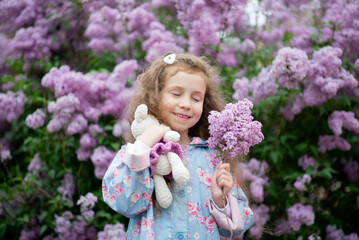 Obraz premium A sweet girl of five years against the background of blooming lilacs enjoys spring and has fun. She has loose long wavy brown hair. Pastel colors. Spring. Childhood. Beauty.