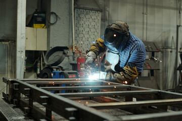 Young male welder in protective mask and gloves welding metallic structure or huge part of industrial machine in workshop