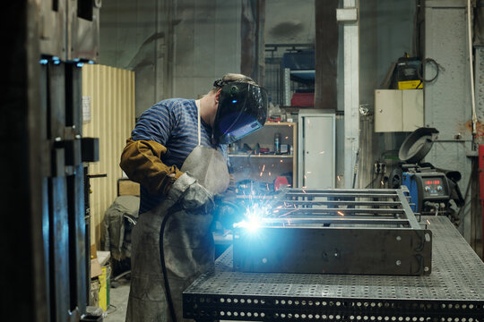Side view of male worker of huge industrial plant or manufactory bending over large metallic workbench while carrying out welding work