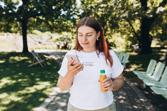 Redhead Girl Walking In The Park And Holding A Bottle Of Cold Smoothie In A Warm Sunny Day. Multivitamin Juice Bottle Mockup. Women Usung Smart Phone. Traveler, Check Social Networks, Send Sms.