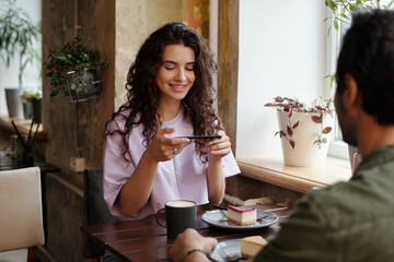 Happy young female with smartphone taking photo of tasty cheesecake on saucer while sitting by table in front of her boyfriend in cafe