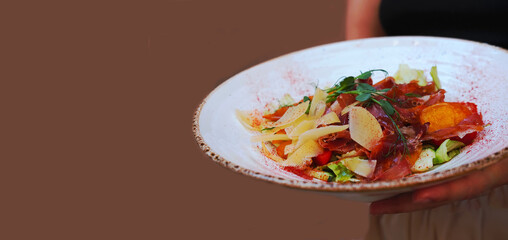 Delicate salad with ripe peach, cherry tomatoes, prosciutto, Parmesan cheese and honey-mustard sauce on beige background. The plate is held by female hands. 