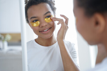Young teen african american girl applying moisturizing under eye patches, looks at mirror. Skincare