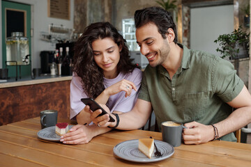 Young couple looking through funny photos or video in smartphone held by guy while having cappuccino or latte with cheesecakes