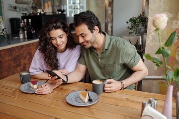 Young couple communicating in video chat in smartphone while sitting by table in cafe and having cappuccino or latte with tasty cheesecakes