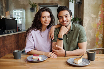 Young cheerful couple looking at camera while sitting by table and having tasty cheesecakes and...