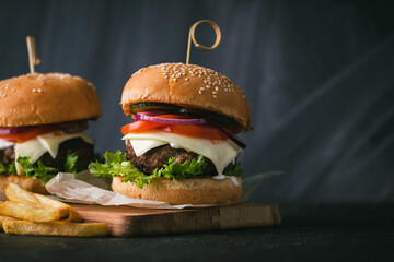 Big beef patty burgers with cheese tomato purple onion cucumber and lettuce next to french fries on wooden black stand on black background