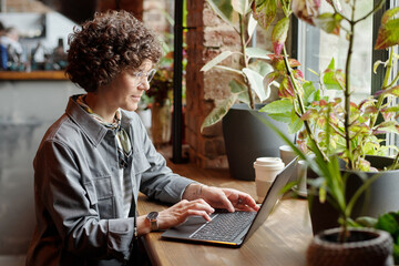 Young serious businesswoman or freelancer in casualwear sitting by window in front of laptop in...