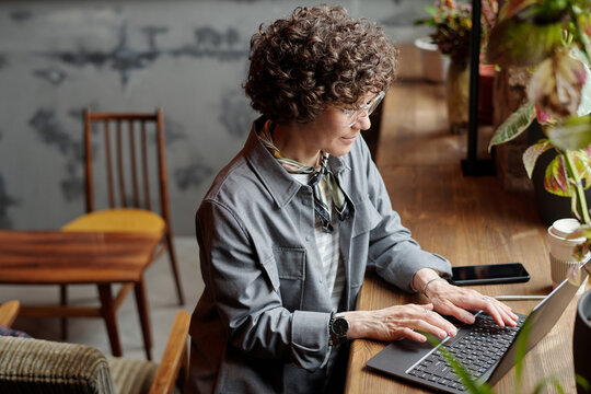 Serious middle aged businesswoman or freelancer with laptop sitting in front of window in cafe, typing on keyboard and looking at screen