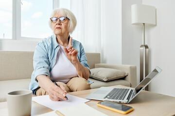 Portrait of an elderly business woman taking notes in a notebook sitting on the couch at home with a laptop undergoing online training