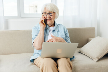 a happy, joyful, pleasant elderly lady is sitting on a cozy sofa in a bright apartment holding a laptop on her lap and happily talking on the phone with her hand leaning against her chest