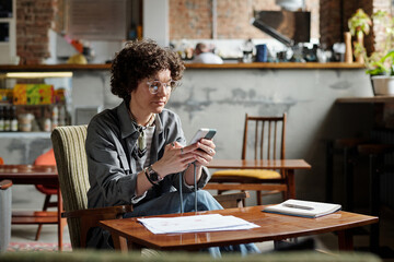 Obraz premium Young female freelancer or manager scrolling in smartphone while sitting in armchair by table with financial documents and notebook in cafe