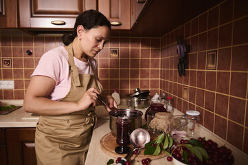 Charming housewife in apron, stands by kitchen countertop and pours freshly made cherry jam into...