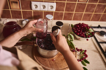 Cropped view of a housewife, woman confectioner pouring delicious freshly brewed cherry berry confiture into sterilized glass jar at home kitchen.