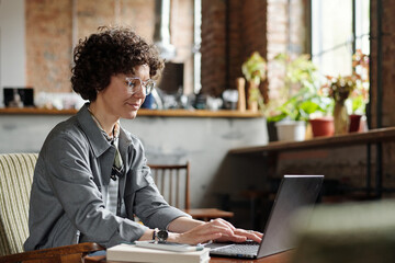 Young female freelancer or owner of cafe in casualwear and eyeglasses sitting in front of laptop by table and organizing work - obrazy, fototapety, plakaty