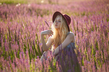 Attractive female model with blonde hair wear hat, posing in a floral field, behind beautiful sunset background.