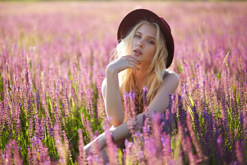 Cheerful female model with blonde hair wear hat, posing in a floral field, behind beautiful sunset background.