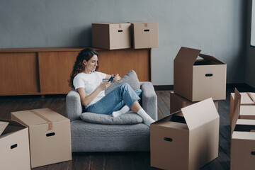 Woman in armchair clicks phone among boxes. European girl having rest in apartment after relocation.