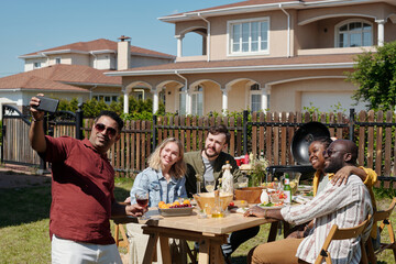 Young cheerful intercultural friends making selfie on smartphone during outdoor dinner or party by table served by homemade food