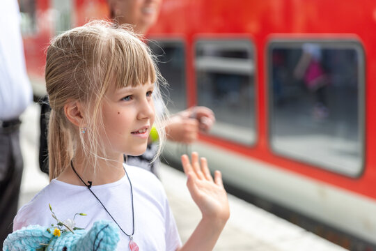 Portait of cute beautiful caucasian little blond kid girl looking waving hand hello goodbye gesture farewell at railway platform station agains red wxpress train. Child meeting friend at railroad