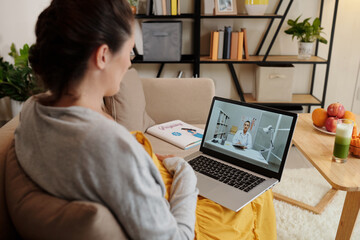Young pregnant woman talking to her doctor online using laptop while sitting on sofa in living room