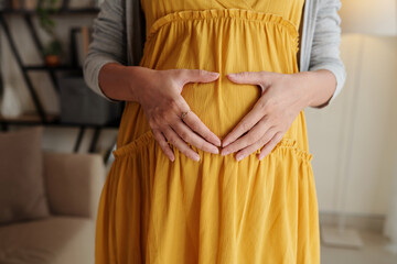 Close-up of young pregnant girl in yellow dress making heart shape with hands on her belly, she expecting baby with love
