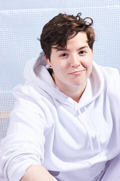 Natural portrait of young handsome transgender man posing with neutral happy face expression. Caucasian with wavy brown hair. Wearing white hoodie and trousers.