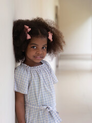 Cute little African-American girl with Afro hair standing against white wall looking at camera.