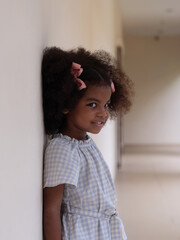Side way image. Cute little African girl with Afro hair standing against white wall smiling with mischievous look.