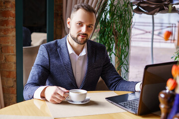 A young man in a business suit sits at a table in a restaurant. Businessman drinking coffee while working at his laptop in a restaurant or cafe