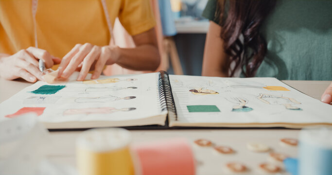 Close-up profession Asia lady designer teamwork meeting sketch book work on new collection share idea in shop. Young tailor discuss color for new cloth in workshop at table, Concept of collaboration.