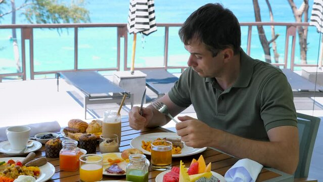 Angry man have breakfast or brunch at restaurant in hotel with turquoise sea background. Sad dissatisfied male tourist at morning in resort cafe. Aggressive, irritated, furious, mad people concept.