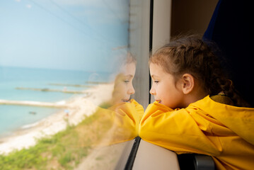 A little girl of 6-7 years old looks out the window of a train at the sea. She's wearing a yellow parka. Journey. Reflection. Vacation. Summer. Family vacation.