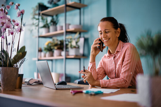 Happy Flower Shop Worker Using Laptop While Talking On Cell Phone At Floral Store.