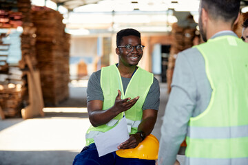 Happy African American worker communicating with inspector at wood warehouse.