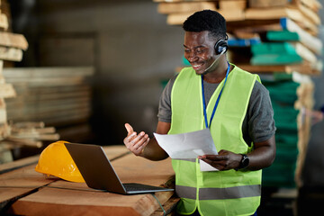 Happy black worker with headset using laptop while having video call at wood warehouse.